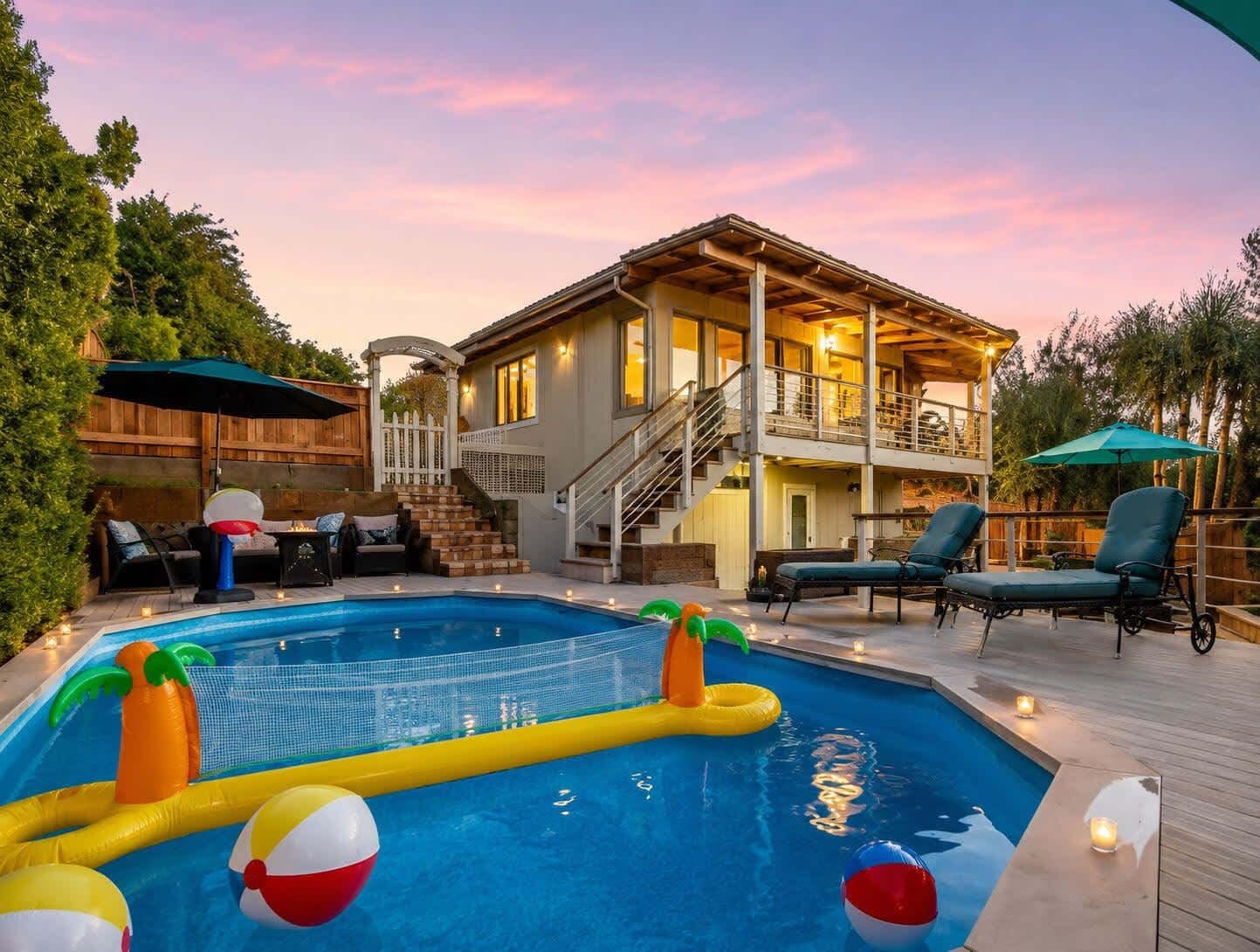 A pool with inflatable toys, a covered deck area, outdoor seating, and a two-story house with a balcony.