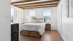 Un dormitorio con estructura de cama de madera, ropa de cama blanca, dos mesitas de noche con lámparas y ventanas amplias con persianas.