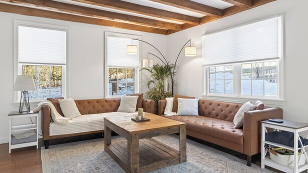 Una sala de estar acogedora con un sofá seccional de cuero marrón, una mesa de centro de madera y una mesita auxiliar blanca. Hay ventanas grandes con persianas blancas, una planta en maceta y una lámpara sobre la mesita.