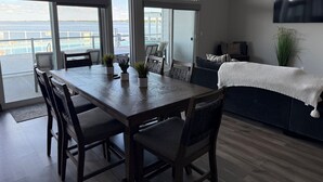 A spacious living area with a wooden dining table, comfortable seating, a sofa, and a view of the sea through large windows. - Spacious 2 bedroom condo on Lake Kampeska (Watertown)