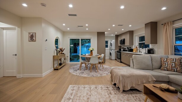 Un salon moderne comprenant un canapé, une table à manger avec des chaises, un coin cuisine équipé d’appareils électroménagers et une vue sur l’extérieur grâce à une porte vitrée.