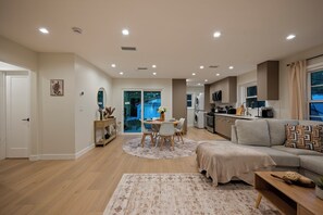 A modern living room with a sofa, a dining table with chairs, a kitchen area with appliances, and a view of the outdoors through a glass door. - Modern 2BR In Woodland Hills EV Charger •Pool•Fast Wi-Fi+WorkDesk•AC Smart-Lock (Los Angeles)
