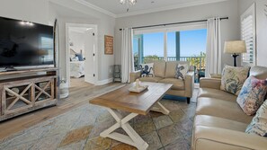 A spacious living room with a large TV, a wooden coffee table, a beige sofa, and a view of the sea through large windows. - Beachfront, Tropical Paradise, Intimate Complex, EV Charger! (Indian Rocks Beach)