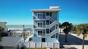 Un edificio de varios pisos con balcones, situado cerca de una playa con palmeras y un cielo despejado.