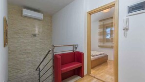 Quarto compacto com um banco vermelho, porta de madeira que dá para um quarto, parede de pedra e unidade de ar condicionado.