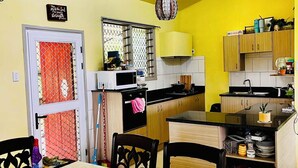 Une cuisine compacte aux murs jaunes, un coin repas avec une table dressée pour deux personnes, et une fenêtre agrémentée d’un rideau décoratif.