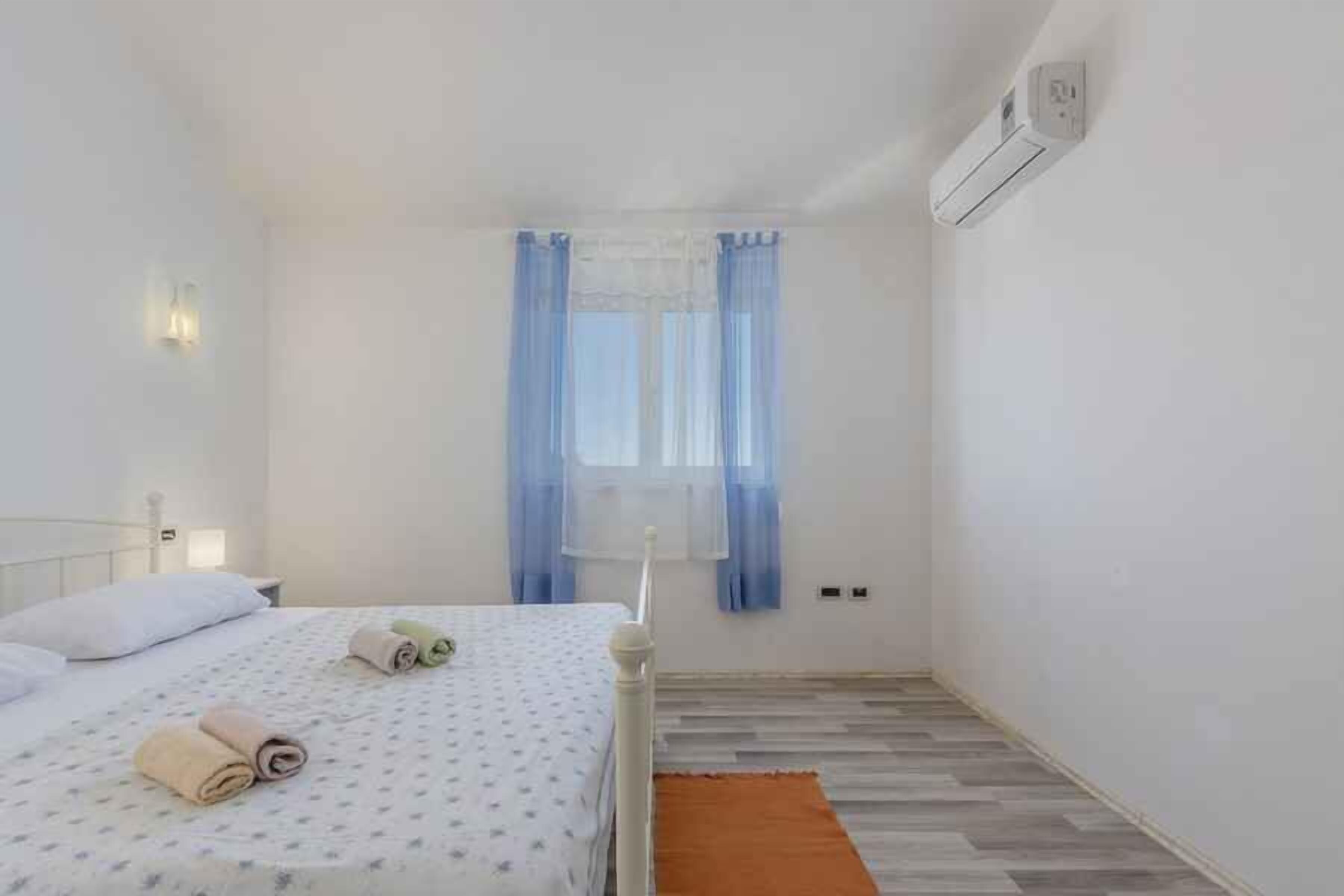 Un dormitorio pequeño y limpio con una cama individual, una ventana con cortinas, aire acondicionado de pared y piso de madera.