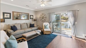A living room with a sofa, armchair, and ottoman, a large mirror, and a view of a lake through glass doors. - Coastal St. Simons Island Townhome Near Beaches, Shopping & Dining (Saint Simons Island)