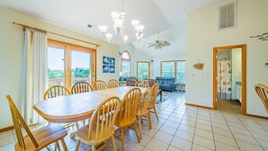 A spacious dining area with a large wooden table and chairs, a chandelier, and large windows offering a view of the outdoors. - Knot A Care - Corolla, Ocean Sands, 400 Yds to Beach, Pool, Hot Tub, Pool Table, Pets OK (Corolla)