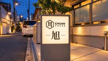 A sign for 'CHUAN HOUSE' with a modern design, located on a street with a parked car and buildings in the background.