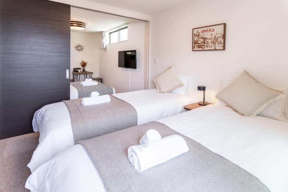 A hotel room with two beds, a TV, a framed picture of Osaka, and a desk with a chair.