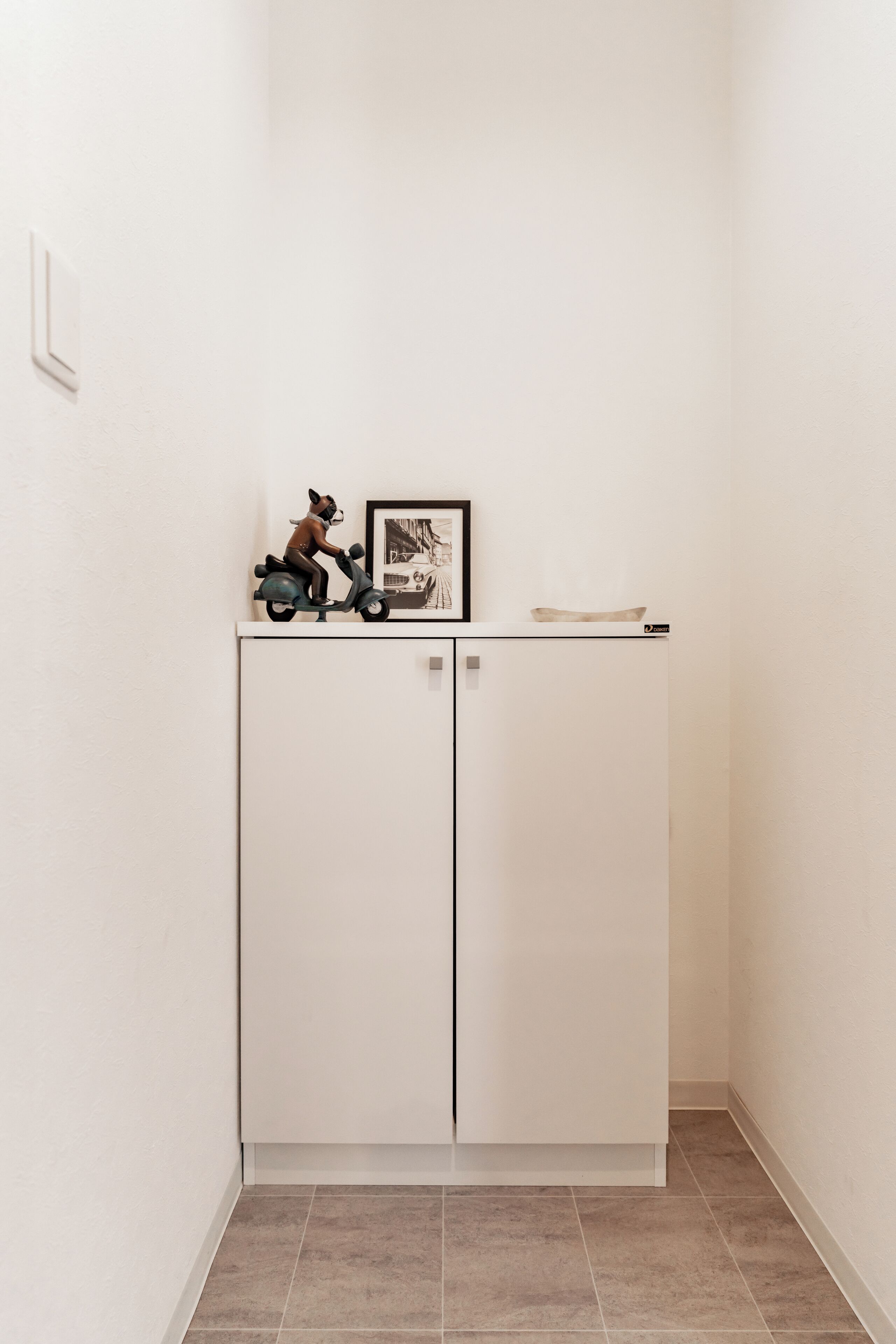 A white cabinet with two doors, a small shelf with a figurine and a framed picture, and a light switch on the wall.
