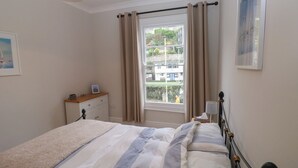 A bedroom with a bed, a bedside table, a lamp, a window with curtains, and a view of buildings outside. - The English School, Flat 2 (Dartmouth)