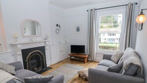 A living room with a fireplace, two grey sofas, a TV on a wooden stand, and a large window with curtains. - The English School, Flat 2 (Dartmouth)