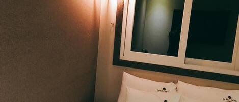 A hotel room with a bed, pillows, a wall-mounted light fixture, and a window.