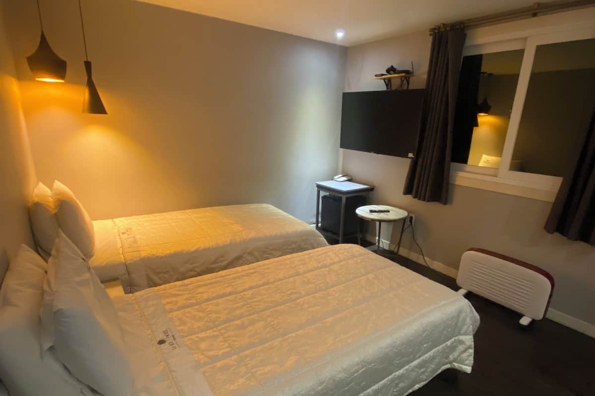 A hotel room with two beds, a small table, a heater, and a window with curtains.