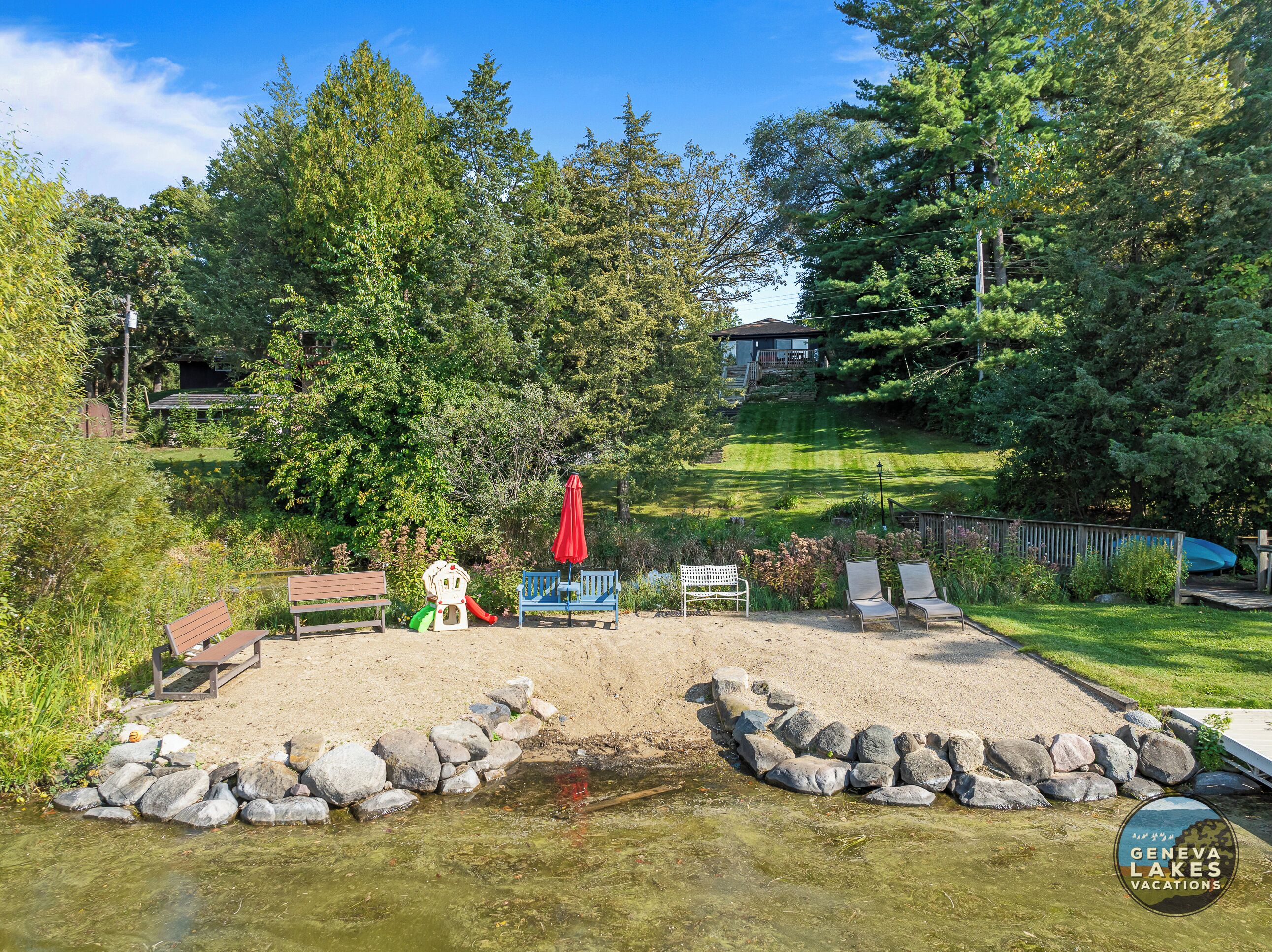 A lakeside park with a sandy play area, benches, and a red umbrella.