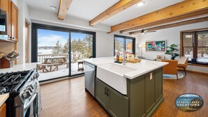 A modern kitchen with a large island, wooden beams, and a view of a snowy deck. - Lakefront Getaway, Swim, Laugh, Family Fun for all! Close to Kettle Moraine (Palmyra)