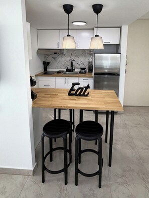A modern kitchen with a wooden table, two black stools, a stainless steel refrigerator, and pendant lights. - Refined apartment with partial ocean-view balcony, steps from Condado Beach. (San Juan)