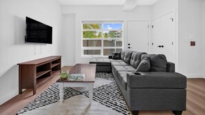 A modern living room with a grey sofa, a wooden coffee table, a flat-screen TV, and a large window with a view of buildings. - Crescent Heights Escape (Calgary)