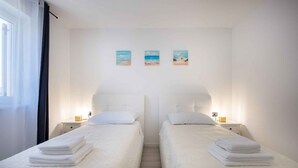 Ein Hotelzimmer mit zwei Betten, jeweils mit weißer Bettdecke und einem ordentlich gefalteten weißen Handtuch darauf. Darüber hängen drei kleine Bilder, die Strandansichten zeigen.