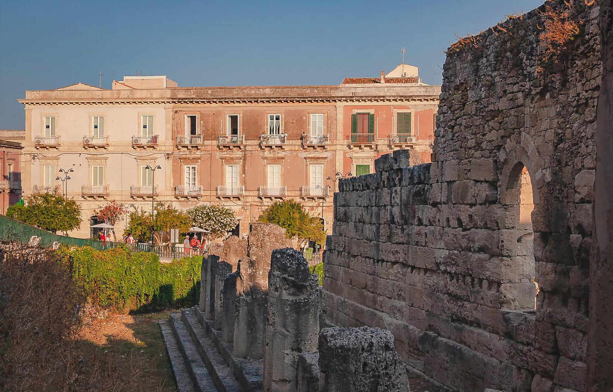 A historical building with classical architecture, featuring a central courtyard with ruins and a large, multi-story structure with balconies in the background.