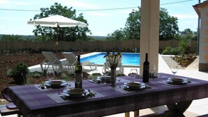 Un área de comedor al aire libre con una mesa preparada para dos personas, una piscina al fondo y un espacio de estar techado con sillas.