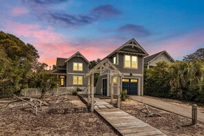 A two-story house with a wooden deck leading to the front door, surrounded by a white picket fence and palm trees. - This like-new 4BR/3.5BA home in sought-after Cape Fear Station- Pet friendly (Bald Head Island)