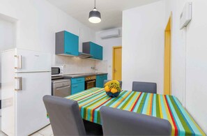 A compact kitchen with a microwave, stove, and white refrigerator. A colorful striped tablecloth covers the dining table, and a fruit bowl sits on it. - Holiday home with pool and air conditioning (Fažana)