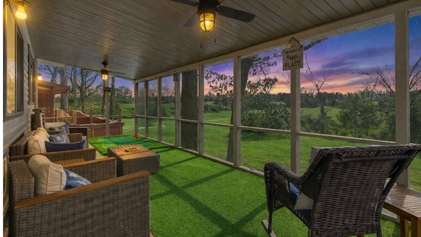 Een overdekt terras met rieten meubilair, een plafondventilator en uitzicht op een zonsondergang over een grasachtig landschap.