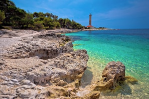 A coastal landscape with rocky terrain, clear turquoise waters, and a lighthouse in the distance. - Holiday apartment with air conditioning (Mali Iž)
