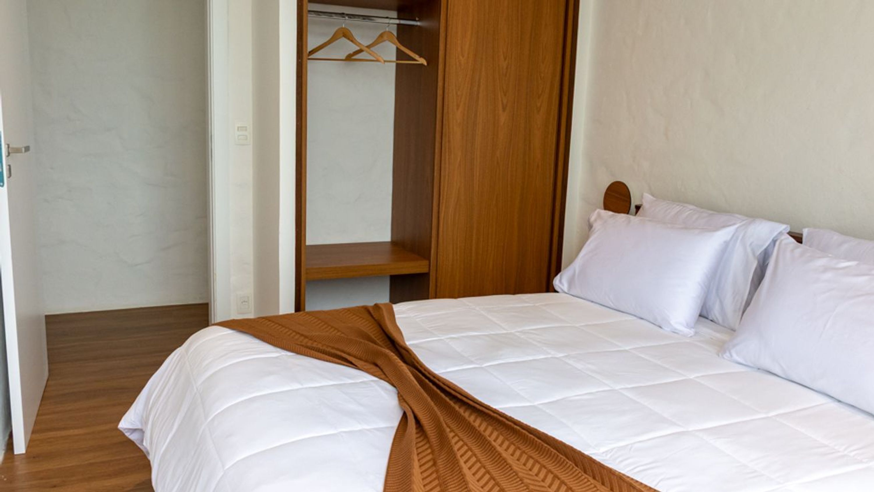 Ein ordentlich bezogenes Bett mit weißen Leinen und einer braunen Tagesdecke, ein hölzernes Kopfende und ein Schrank mit Kleiderstange.