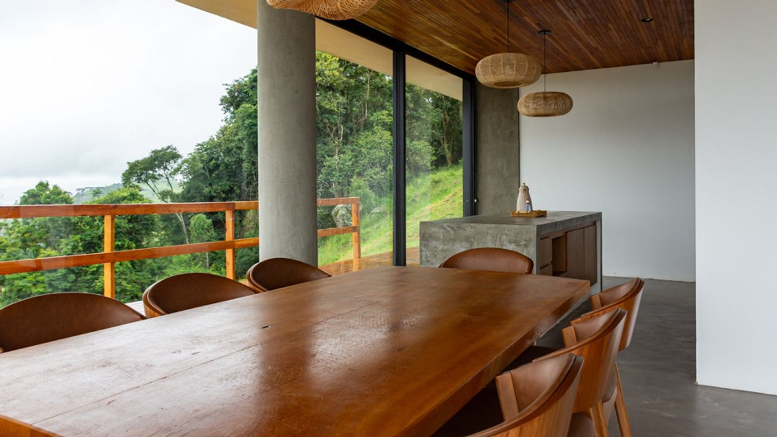 A dining area with a long wooden table and chairs, a small kitchen counter, and large windows offering a view of a lush landscape.