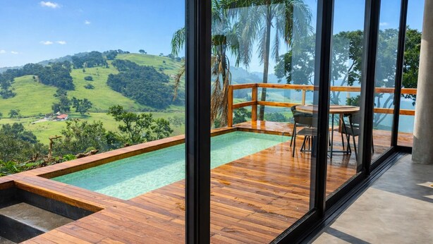 A wooden deck with a pool, a table, and chairs, overlooking a lush landscape with hills and trees. - Casa c Piscina e Vista Panor Mica Bueno Brand o (Pouso Alegre)