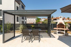 Eine Terrasse im Freien mit überdachter Sitzgelegenheit, einem Grill und Blick auf benachbarte Gebäude.