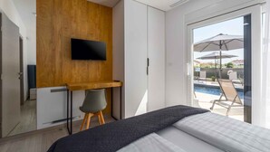 A bedroom with a bed, a desk, a TV, and a view of a pool and outdoor seating. - Holiday apartment with air conditioning and shared pool (Funtana)