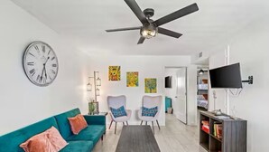 Un soggiorno moderno con un divano color petrolio, due poltrone, un tavolino da caffè, una ventola a soffitto, una sveglia a parete e un televisore montato al muro.