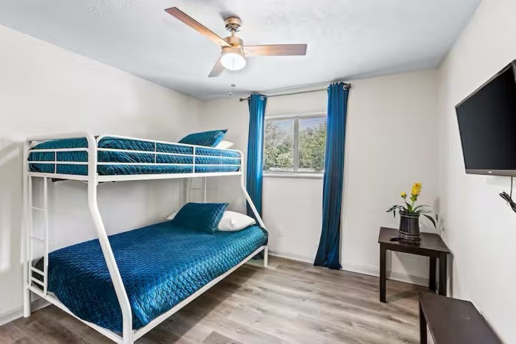 A bunk bed room with a ceiling fan, a window with curtains, a small table with a plant, and a TV on the wall.