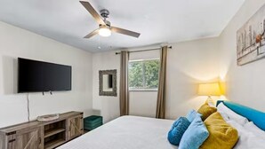 Una camera da letto con un letto, una ventola a soffitto, una TV a schermo piatto, una finestra con tende e un quadro appeso al muro.