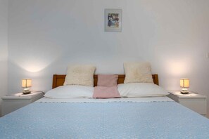 Ein ordentlich bezogenes Bett mit einer blauen, gemusterten Tagesdecke, zwei weißen Kissen und einer pinkfarbenen Zudecke. Auf weißen Nachttischen stehen jeweils zwei Nachttischlampen.