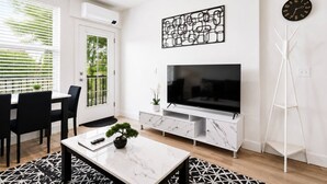 Een moderne woonkamer met een flatscreen televisie, een witte marmeren salontafel, een eethoek met stoelen en een groot raam met jaloezieën.