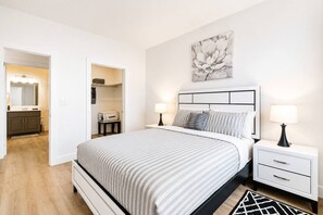 Une chambre à coucher avec un lit, des tables de chevet, une commode, une lampe, un tableau et une salle de bain visible à travers une porte ouverte.