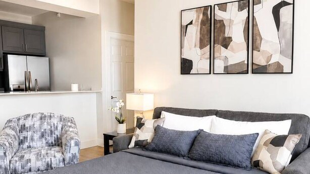 Een slaapkamer met een groot bed, een grijze bank en een keuken in de verte.