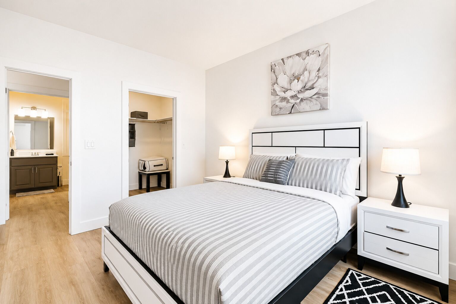 A bedroom with a bed, bedside tables, a nightstand, a lamp, a painting, and a bathroom visible through an open door.