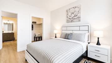 A bedroom with a bed, bedside tables, a nightstand, a lamp, a painting, and a bathroom visible through an open door.
