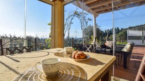 Ein Tisch mit Tasse, Untertasse und Croissant auf einer sonnigen Terrasse mit Blick auf Bäume und einen weißen Zaun.