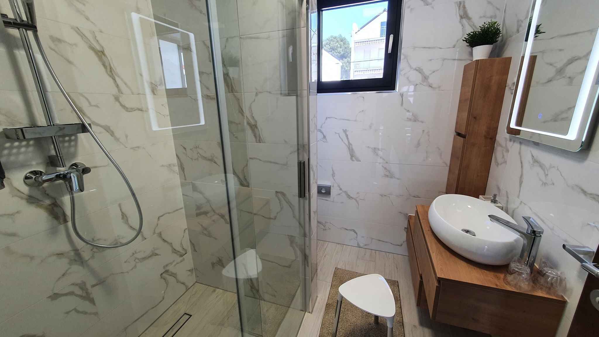 Un bany modern amb plat de dutxa de vidre, un tocador de fusta i un lavabo blanc.