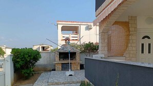 A stone-tiled patio with a barbecue area, a small garden with plants, and a building with a tiled roof and a balcony. - Holiday apartment with barbecue facilities and terrace (Vir)