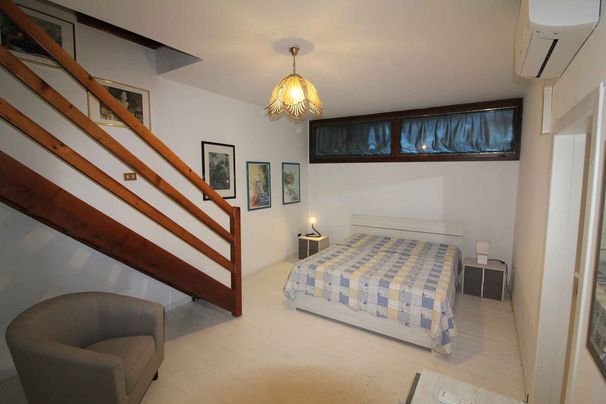 Une chambre avec un escalier en bois, un lit recouvert d’une couverture à carreaux, une table de chevet avec une lampe et une fenêtre avec des rideaux bleus.