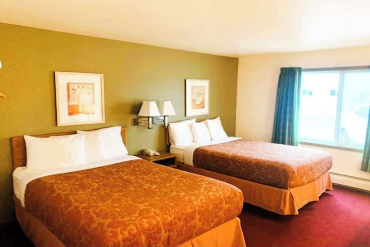 A hotel room with two beds, a nightstand with lamps, framed artwork on the wall, and a window with curtains.